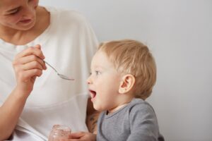 Young attractive mother with her one years old little son dressed in pajamas giving him his first fruit smoothies food in the bedroom at the weekend together, lazy morning.