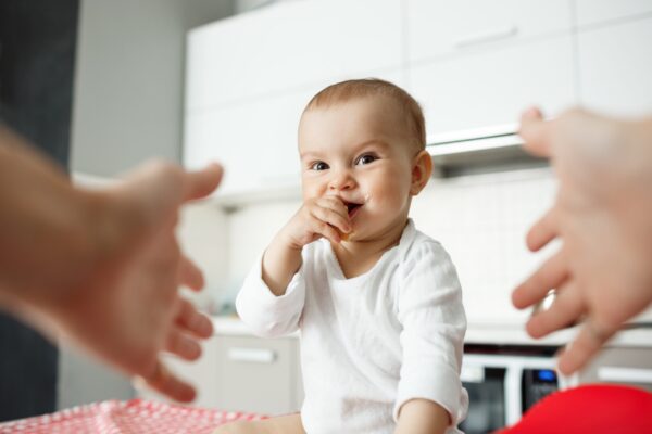 Close up portrait of happy little baby boy with big eyes putting hand in mouth and smiling brightdully. Mother hands in frame