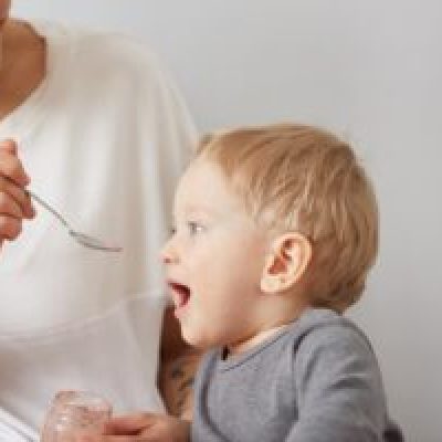 Young attractive mother with her one years old little son dressed in pajamas  giving him his first fruit smoothies food in the bedroom at the weekend together, lazy morning.