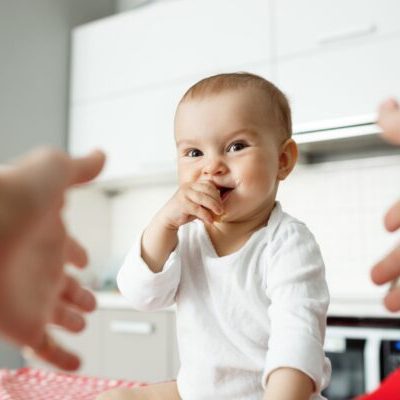 Close up portrait of happy little baby boy with big eyes putting hand in mouth and smiling brightdully. Mother hands in frame